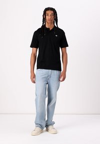 Svart polo skjorte med korte ermer og logo på venstre bryst, sammen med lyseblå rette jeans og kremfargede joggesko.