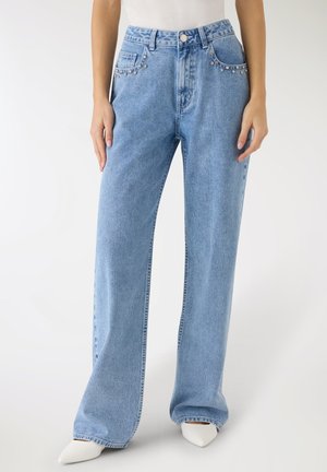 Donna che indossa jeans a vita alta e gambe larghe di colore azzurro chiaro con borchie piccole d'argento sulle tasche anteriori e scarpe bianche con punta affilata.