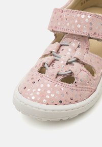 Rosa semsket sandal med skinnende sølv- og rosa prikkede mønstre, et åpent design med grå elastiske snorer, og en hvit gummisåle.
