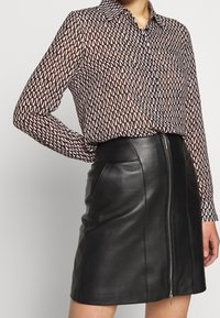 Vrouw in een zwarte leren rok met rits aan de voorkant en een lange mouwen blouse met patroon, gestoken in de rok, staande met één hand op de heup.