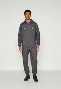 Mørk grå treningsdrakt bestående av en langermet pullover med krage, som har blå detaljer og gulllogo, sammen med matchende smale bukser.