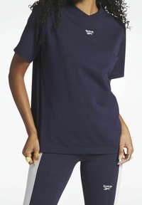 Námořnická modrá tričko s krátkým rukávem a výstřihem do V s logem Reebok. Nosené s odpovídajícími legínami s bílým bočním pruh a těsným střihem.