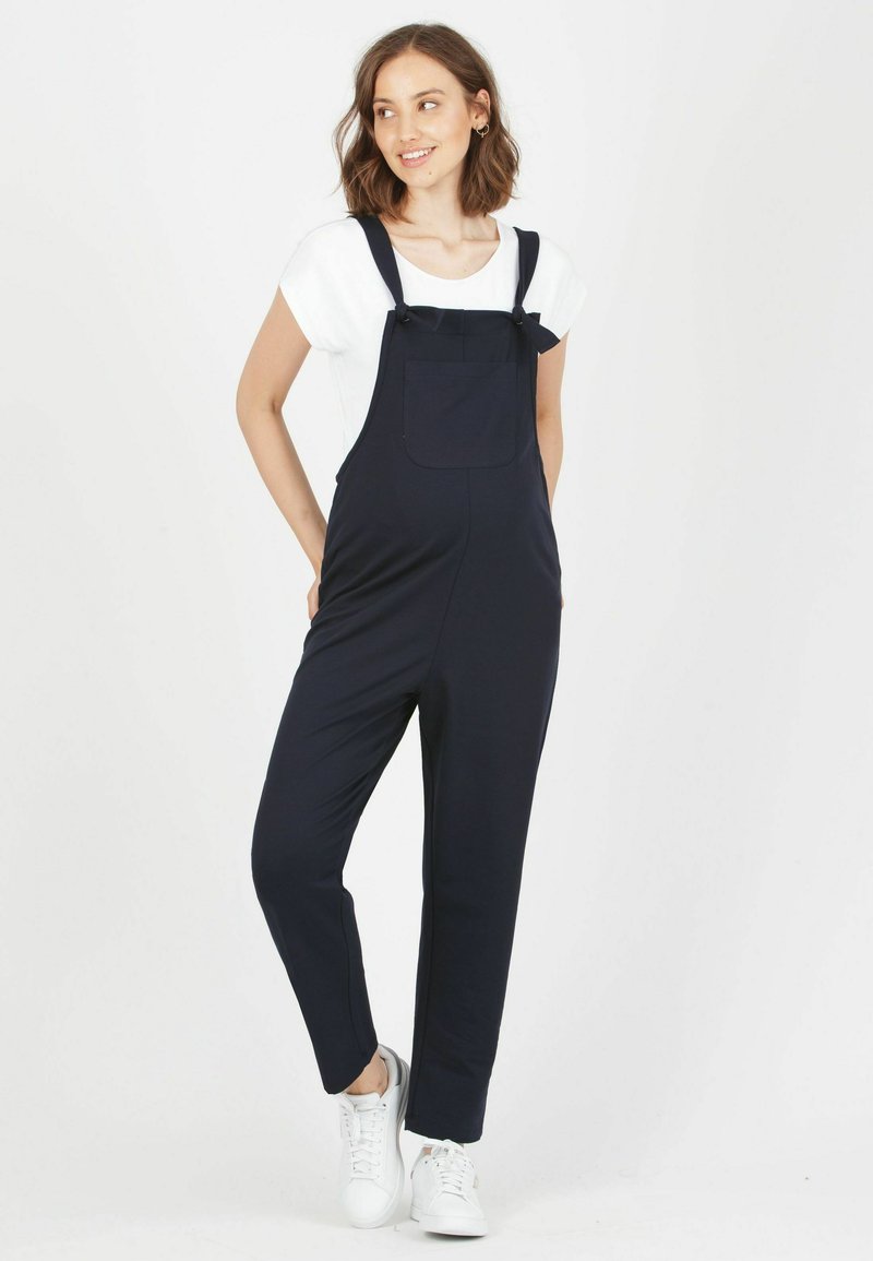 Mörkblå overaller med justerbara axelremmar, en framficka och rak benstil, bärs över en vit T-shirt med vita sneakers.