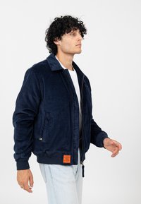 Marineblauwe corduroy jas met een rits aan de voorkant, kraag, zijzakken en gebreide manchetten. Kenmerkt zich door een opvallend oranje merklabel aan de zoom.