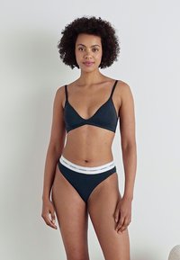 Fekete bralette vékony pántokkal és hozzá illő, magas derekú tangával, amelyen fehér, elasztikus derékszíj található a "GUESS" felirattal. Sima szövetű textúra.