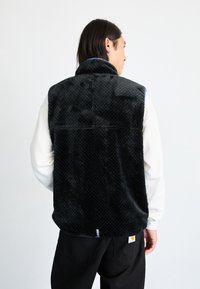 Black fleece vest with a textured pattern, high collar, and open back. Worn over a white long-sleeve shirt, featuring a logo patch at the hem.
