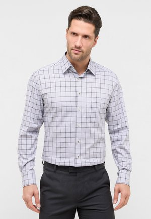 Mand iført lysegrå ternet langærmet skjorte med knapper og mørke bukser, stående mod en ensfarvet hvid baggrund.