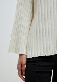 White ribbed knit top with flared sleeves, featuring a raw hem and soft texture, paired with black fitted pants.