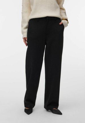 Personne portant un pantalon noir ample, un pull en maille crème, des chaussures noires pointues, avec une main dans la poche et l'autre détendue sur le côté.