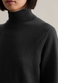 Rövid sötét hajú nő fekete garbós pulóvert visel, látható a nyaka, válla, és az ajkai zárva vannak egy semleges háttér előtt.