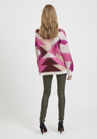 Maglione over size multicolore con motivi geometrici in rosa, crema e marrone; abbinato a pantaloni verdi oliva aderenti e tacchi neri.