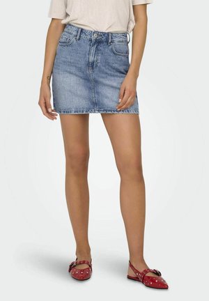 Femme portant un haut beige clair, une jupe courte en jean bleu et des chaussures plates rouges cloutées, debout devant un fond uni.