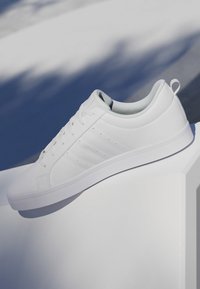 Zapatilla blanca de caña baja con cordones, tres franjas laterales y un lazo en el talón, exhibida sobre una superficie blanca con fondo sombreado.