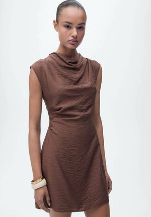 Brun cowl-neck klänning med korta ärmar, texturerad tyg och åtsittande design. Bärs med ett vitt armband på handleden, modellen vänder sig framåt.