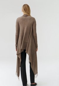 Larga chaqueta de punto abierta en marrón tierra con un dobladillo trasero drapeado y detalles de flecos. Llevada sobre pantalones negros, con la textura del tejido visible.