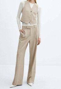 Beżowy zestaw trzyczęściowy składający się z dopasowanej kamizelki, bluzki z długim rękawem oraz spodni z szerokimi nogawkami, wykonany z gładkiego materiału. Wyraziste białe szpilki w kształcie szpicy.