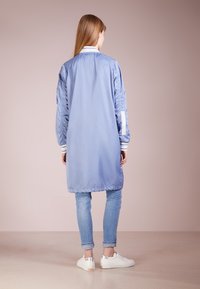 Chaqueta larga y ligera en nylon azul claro con cuello acanalado, puños elásticos y finas franjas laterales; llevada sobre pantalones de mezclilla y zapatos blancos.