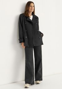 Dubbelgebreide zwarte peacoat met grote knopen, gedragen over een wit shirt. Gecombineerd met donkere flared jeans en beige sneakers. Zachte textuur.