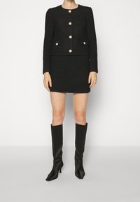 Zwart wollen blend jasje en mini-jurk set, met goudkleurige knoopversieringen, gecombineerd met kniehoge zwarte stiletto laarzen.
