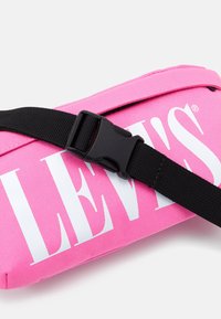 Różowa torba na ramię z białym logo, z teksturowaną powierzchnią, regulowanym czarnym paskiem i plastikowym zapięciem.