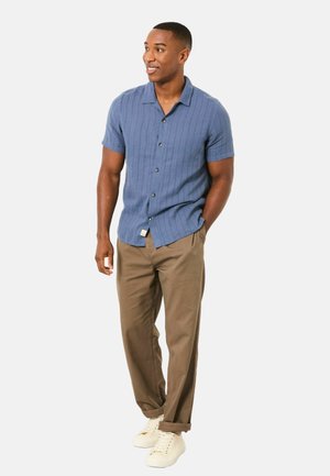 Kurzärmeliges blaues Hemd mit Knöpfen und einer strukturierten Musterung, kombiniert mit beigefarbenen Hosen und weißen Turnschuhen. Modell steht in entspannter Haltung.