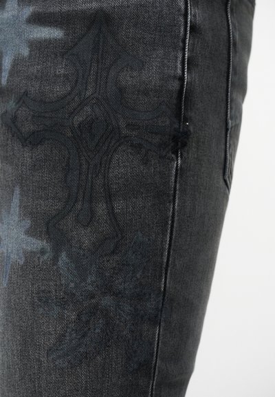 Κοντινό πλάνο σκούρου γκρι τζιν με αχνή μαύρη κεντητή διακόσμηση σε σχήμα σταυρού στο μηρό και εμφανείς ραφές στη πλευρά.