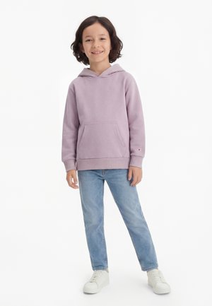 Bambino sorridente con capelli corti e scuri che indossa una felpa con cappuccio viola chiaro, jeans blu e scarpe da ginnastica bianche, in piedi su uno sfondo bianco.