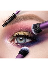 Gros plan d'un œil bleu avec fard à paupières doré et violet, sourcil coiffé avec un spoolie, et pinceaux appliquant du maquillage autour de l'œil.