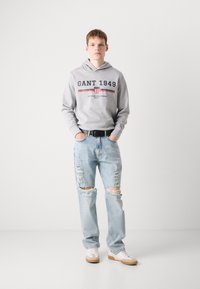 Jeune homme portant un sweat à capuche gris "GANT 1949", un jean déchiré bleu clair, une ceinture noire et des baskets blanches, se tenant devant un fond blanc.