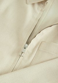 Tejido beige con un tejido texturizado, que presenta un cierre metálico y un forro interior suave. Incluye un bolsillo de parche cerca del cierre.