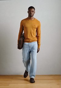 Brązowa skórzana torba na ramię, musztardowy sweter z długim rękawem, jasnoniebieskie luźne dżinsy, ciemne buty; model idący na tle neutralnym.