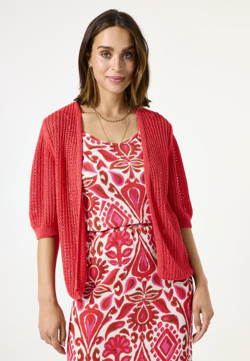 Cardigan rouge tricoté à manches courtes et bouffantes, associé à une robe à motifs floraux avec des teintes de rose, rouge et marron.
