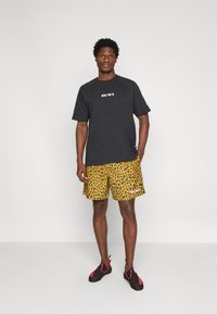 Black short-sleeve shirt with logo, paired with yellow leopard print shorts. Black climbing shoes with red accents on the sole.