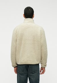 Muškarac u bež flis jakni s visokim ovratnikom, prikazan odostraga, u kombinaciji s tamnoplavim trapericama na jednostavnoj pozadini.