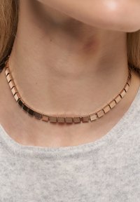 Chokerhalsband med reflekterande fyrkantiga plattor i bruna nyanser, bärs runt halsen ovanpå en ljusgrå texturerad topp.