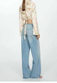 Femme portant un haut cropped à manches longues fleuri noué dans le dos, associé à un pantalon large en denim bleu clair et des sandales.