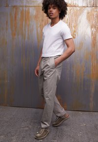 White v-neck t-shirt, beige checkered trousers with side pockets, and patterned olive-green sneakers, standing against a textured metallic background.