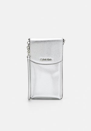 Bolso bandolera plateado con cierre de solapa, logotipo de Calvin Klein y detalle de correa de eslabones sobre un fondo liso.