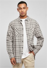 Grijze geruite flanellen shirt met zwarte en witte accenten, twee voorzakken, knoopsluiting, gedragen over een wit T-shirt en lichte broek.