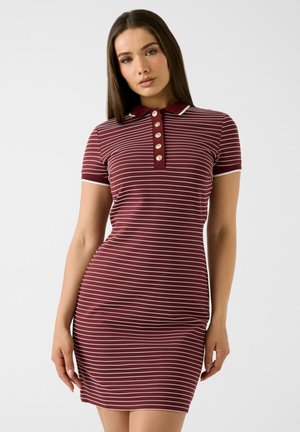 Femme portant une robe bordeaux à manches courtes avec des rayures blanches horizontales et un col boutonné, se tenant devant un fond uni.