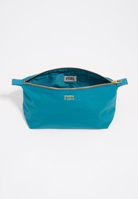 Funda de lona en color verde azulado con un zipper dorado y asa, que presenta una etiqueta con el logo y una superficie lisa y mate. El interior está forrado con tela a juego.