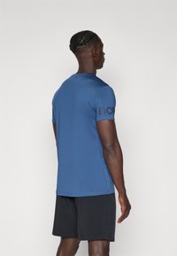 Blå kortermet skjorte laget av glatt stoff, med rund hals og en stor svart logo på venstre erme. Svart shorts kompletterer antrekket.