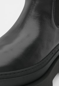 Černá kožená kotníková bota s lesklým povrchem, vybavená robustní podrážkou a elastickými bočními panely pro snadné obouvání. Unikátní prošívání podél podrážky.