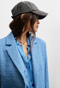 Grauem Hut mit gebogenem Schirm, strukturierte blaue Jacke mit einer unebenen Oberfläche und eine hellblaue Bluse mit floralen Akzenten. Perlenkette sichtbar.