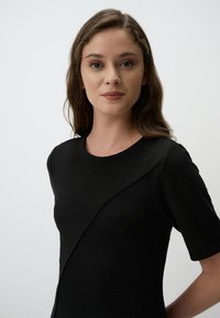 Robe noire texturée à manches courtes, avec un col rond et un détail de couture en diagonale sur le devant. Tissu lisse avec un motif subtil.