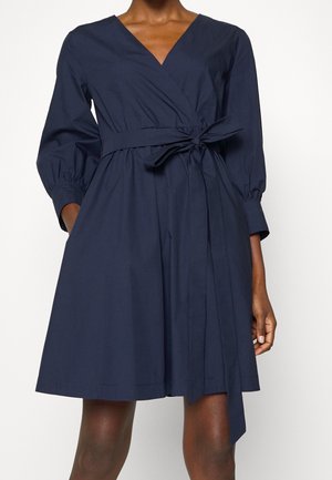 Vrouw draagt een marineblauwe wikkeljurk met lange pofmouwen en een strikceintuur in de taille, staand tegen een effen achtergrond.