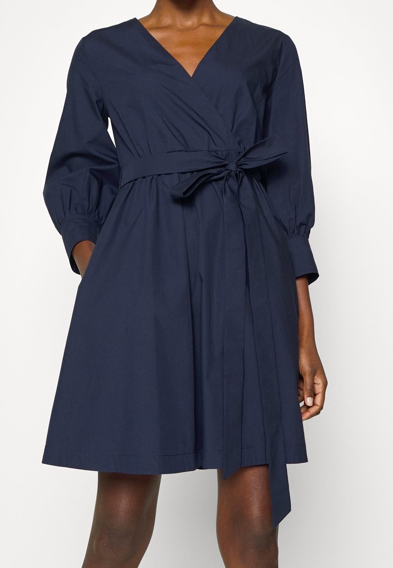 Kobieta ubrana w granatową sukienkę kopertową z długimi bufkami i przewiązanym paskiem w talii, stojąca na jednolitym tle.