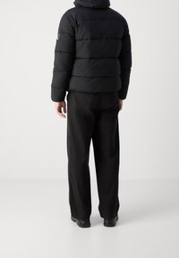 Doudoune noire matelassée horizontalement avec capuche, associée à un pantalon large noir. Logo visible sur la manche de la veste.