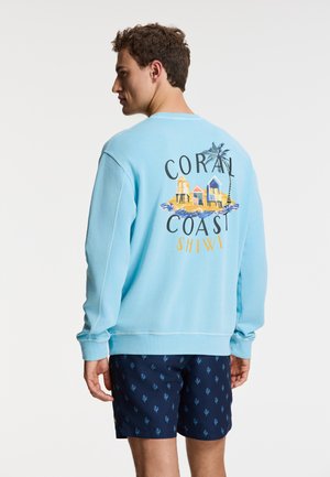 Felpa azzurra con grafica sul retro che recita "CORAL COAST SHIWI," abbinata a pantaloni corti blu scuro con un piccolo motivo blu.