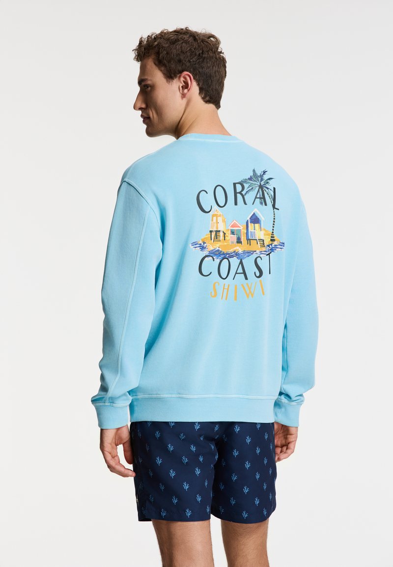 Felpa azzurra con grafica sul retro che recita "CORAL COAST SHIWI," abbinata a pantaloni corti blu scuro con un piccolo motivo blu.